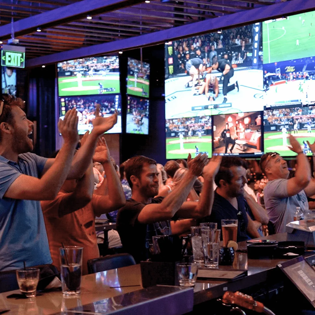 Guests cheering beneath game screens at Tom’s Watch Bar Indianapolis. Source: https://tomswatchbar.com/wp-content/uploads/2025/02/IND-6.png