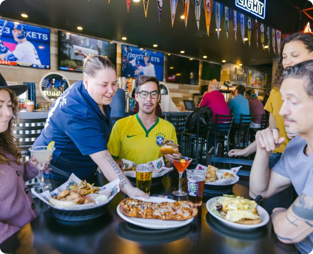 Clientes en una mesa con banderas deportivas y televisores al fondo en Bout Time West Jordan Source: https://www.bouttimepub.com/wp-content/uploads/2022/11/Rectangle-36-1024x831.jpg