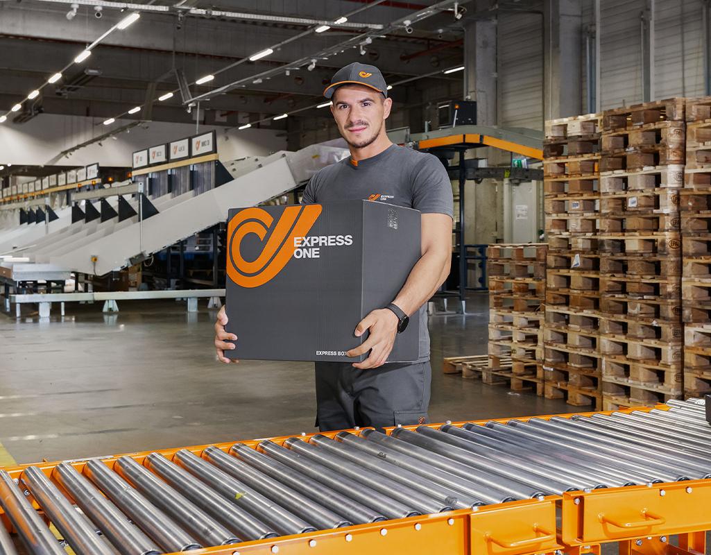 Express One worker holding a parcel beside a conveyor belt at a distribution facility.