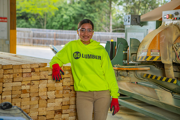 Empleado de 84 Lumber trabaja junto a una sierra y madera apilada / 84 Lumber employee working beside a saw and stacked lumber