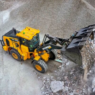 Cargador amarillo trabaja sobre una superficie de grava junto a un contenedor. / Yellow loader works on a gravel surface beside a skip.