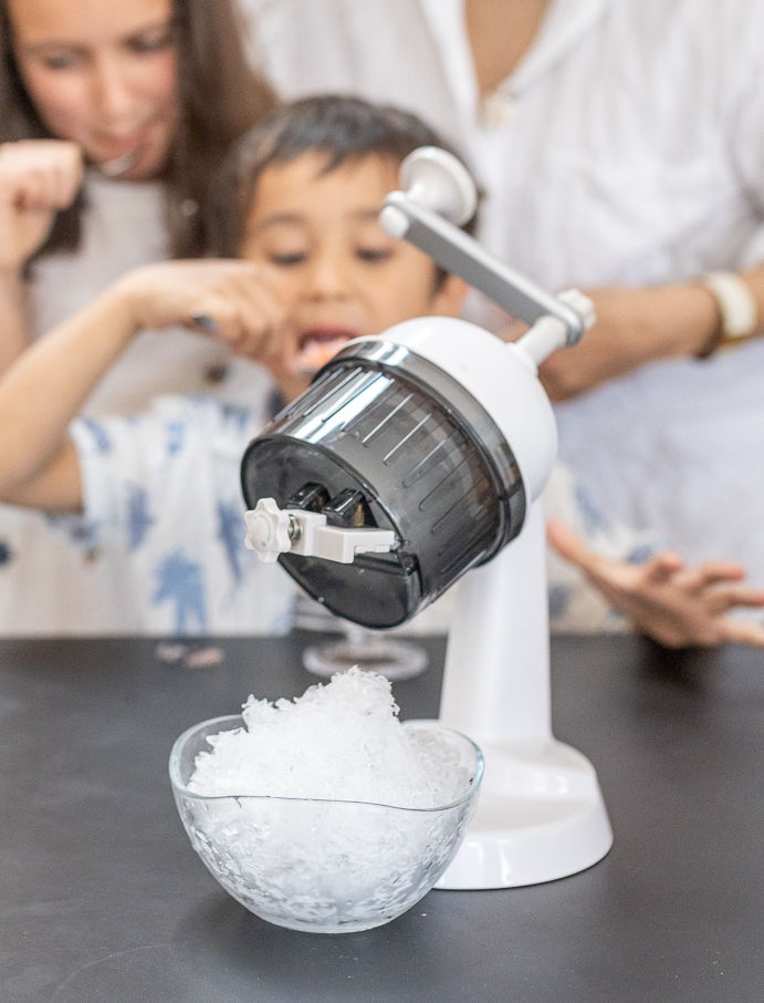 Una persona acciona una máquina Snowpop mientras otra observa cómo cae el hielo raspado en un cuenco. / One person turns a Snowpop machine while another watches shaved ice fall into a bowl.