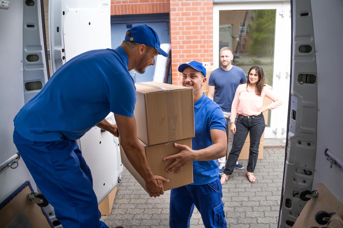 Equipo de mudanza carga una caja junto a un camión y una vivienda / Moving crew loads a box beside a truck and home